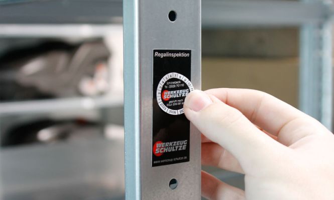 Plakette zur Regalinspektion wird an Metallregal angebracht – dokumentierte Prüfung für sichere und vorschriftskonforme Lagertechnik.